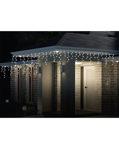 White LED curtain string lights draped over the eaves and columns of a house porch at night, casting a warm glow.
