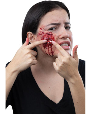 Woman with a bloody wound on her face, pointing at it with both hands.