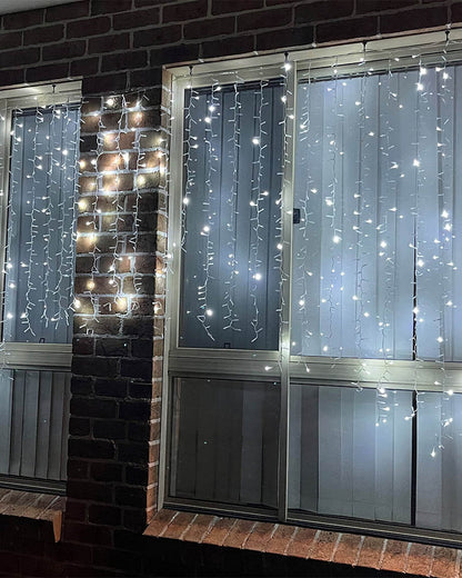 White curtain string lights draped vertically across large windows on a brick building exterior at night.