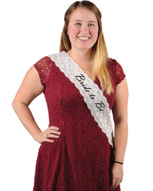 A woman wearing a red dress and a white lace sash with the text 'Bride To Be' on it.
