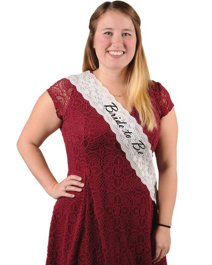 A woman wearing a red dress and a white lace sash with the text 'Bride To Be' on it.